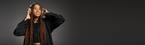 joyful and young African American girl in 20s with headphones listening to music on grey, banner