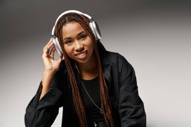 pleased african american woman in 20s with dreadlocks listening music in headphones in grey studio clipart