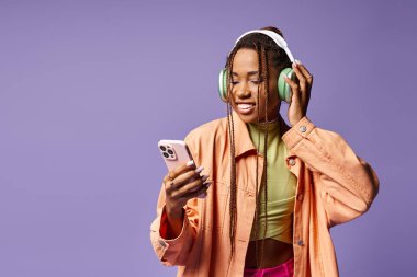 Mutlu Afro-Amerikalı kadın kulaklıklarıyla müzik dinliyor ve eflatun fon üzerinde telefon kullanıyor.