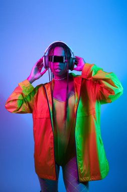 Kablosuz kulaklıklı ve ceketli genç Afrikalı Amerikalı kadın stüdyoda neon ışıklarıyla bekliyor.