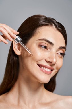 happy young woman with brunette hair smiles as she applying serum with cosmetic pipette in a studio setting. clipart