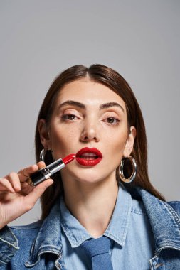 A young Caucasian woman with brunette hair gracefully applying lipstick to her lips in a studio setting. clipart