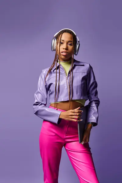 pretty african american woman with headphones holding laptop and standing on purple background
