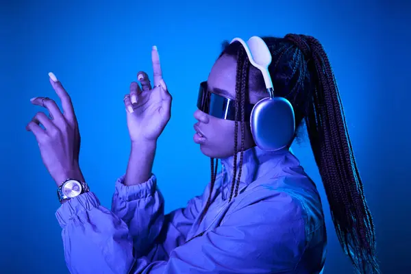 stock image dreamy african american woman posing in trendy sunglasses with headphones on blue with neon light