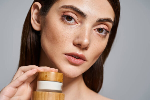 A young Caucasian woman with brunette hair delicately holds a jar of cream in her hand, showcasing her radiant beauty.