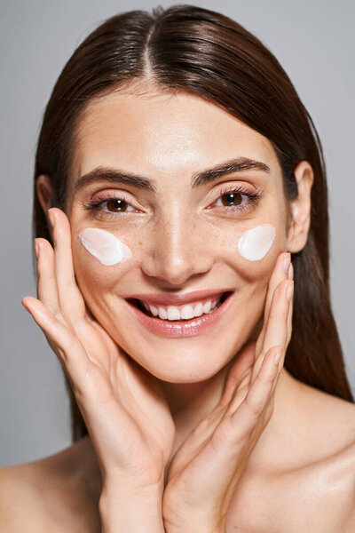 Молодая счастливая женщина с брюнетками и чистой кожей радостно носит толстый слой крема на лице.