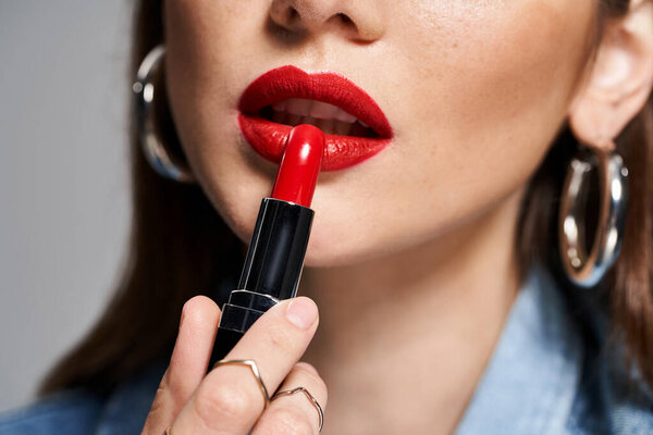 A young Caucasian woman with brunette hair applying bright red lipstick to her lips in a studio setting.