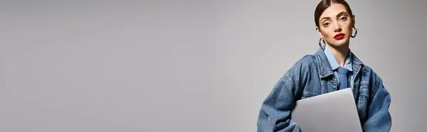 stock image A young Caucasian woman with brunette hair wearing a jean jacket, holding a laptop in a studio setting, banner