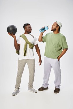 happy african american man holding ball and looking at friend drinking water on grey background clipart