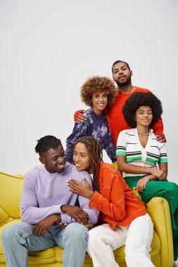 Mutlu Afro-Amerikan arkadaşları, parlak günlük kıyafetleriyle birlikte gri koltukta oturuyorlar.