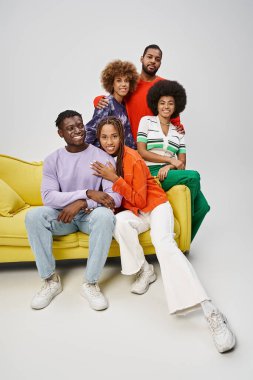 Parlak günlük giysiler içinde mutlu Afro-Amerikan insanları gri koltukta birlikte oturuyorlar.