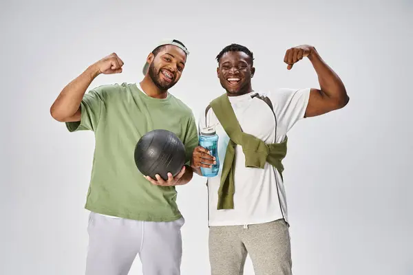 Juneteenth and sport, happy african american men holding ball and water bottle on grey backdrop