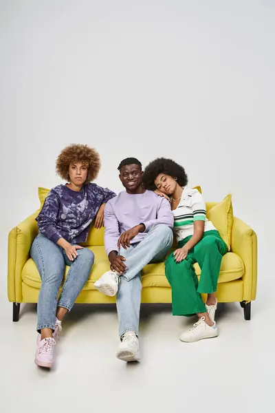 cheerful and curly african american friends together sitting on yellow sofa on grey background