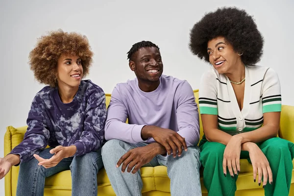 happy and curly african american friends chatting and sitting on yellow sofa on grey background