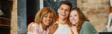 carefree and trendy multiethnic friends embracing and looking at camera in youth hostel, banner clipart