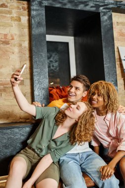 youthful woman taking photo with joyful multicultural friends in lounge of modern students hostel clipart