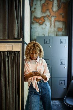 Akıllı telefonlu mutlu Afro-Amerikan kadın yurttaki çift katlı yatakların yanındaki kameraya bakıyor.