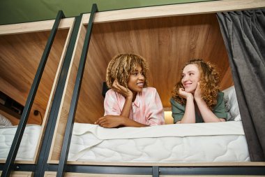 joyful multicultural girlfriends talking on double-decker bed in comfortable students hostel clipart