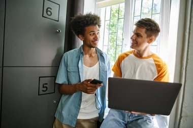 Akıllı telefonlu neşeli Afrikalı Amerikalı öğrenci yurttaki dolabın yanında dizüstü bilgisayar kullanıyor.