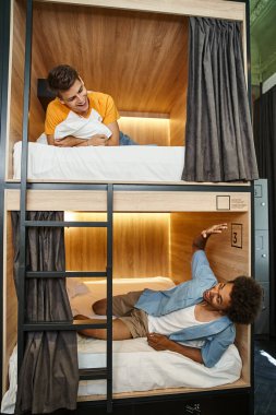 Heyecanlı Afrikalı Amerikalı öğrenci çift katlı yatakta uzanıyor ve otel odasında el sallıyor.