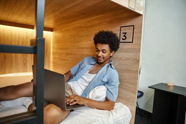 Mutlu ve stil sahibi Afrikalı Amerikalı öğrenci yurttaki çift katlı yataklarda dizüstü bilgisayarla iletişim kuruyor.