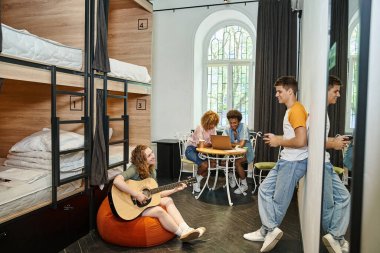 multiethnic buddies with guitar and laptop in room of cozy youth hostel, friendship and travel clipart