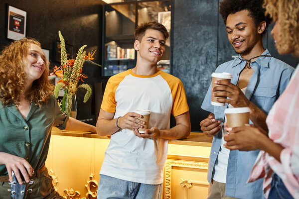 joyous multiethnic buddies with takeaway drinks talking at reception desk in youth hostel, travel