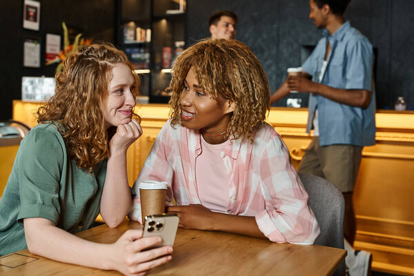 joyful interracial female friends with smartphone and coffee to go talking in lobby cafe of hostel