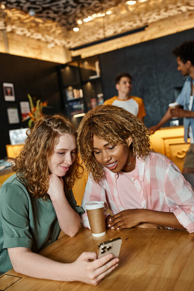 happy woman browsing social media on smartphone near african american female friend in lobby cafe