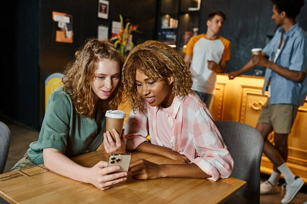 interracial girlfriends browsing internet on smartphone near coffee to go in cafe of hostel lounge