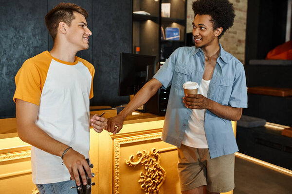 cheerful african american man with paper cup talking to smiling friend at reception desk in hostel