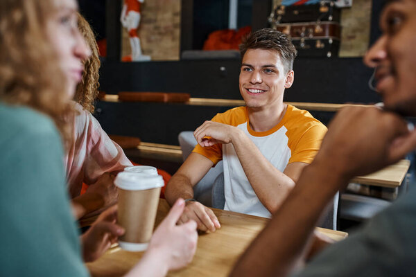 blurred woman with paper cup talking to multiethnic friends in lounge cafe of hostel, fun and travel