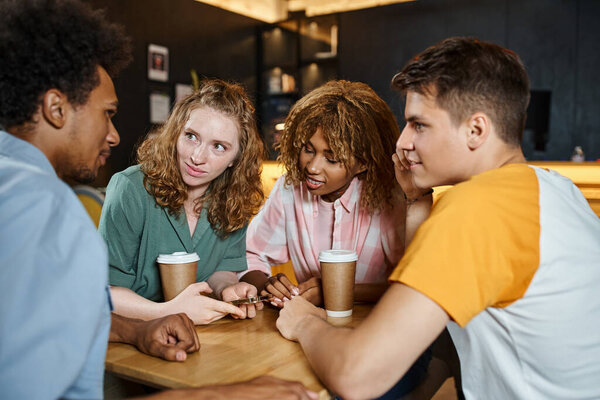 young and stylish multiethnic friends talking near takeaway drinks in lobby cafe of students hostel