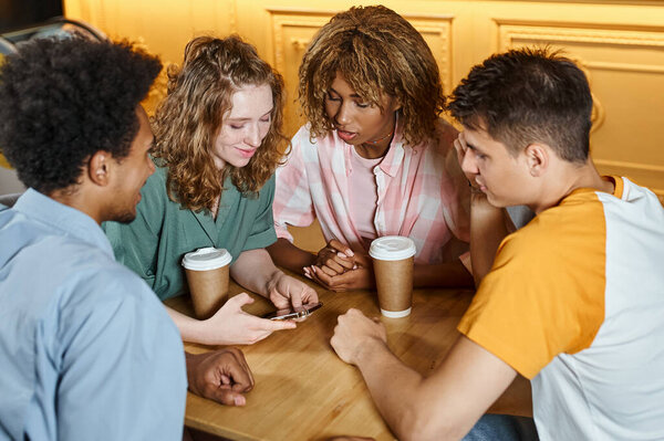 smiling woman browsing internet on smartphone near interracial buddies in cafe of youth hostel