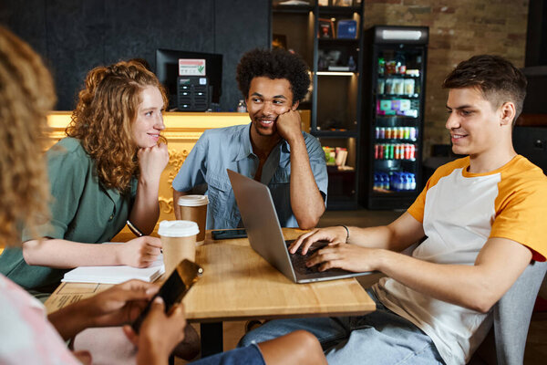 cheerful multiethnic friends talking near student networking on laptop in lounge of youth hostel