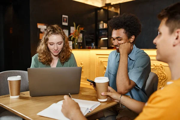 stock image young smiling woman using laptop near multiethnic men with smartphone and notebook in hostel cafe