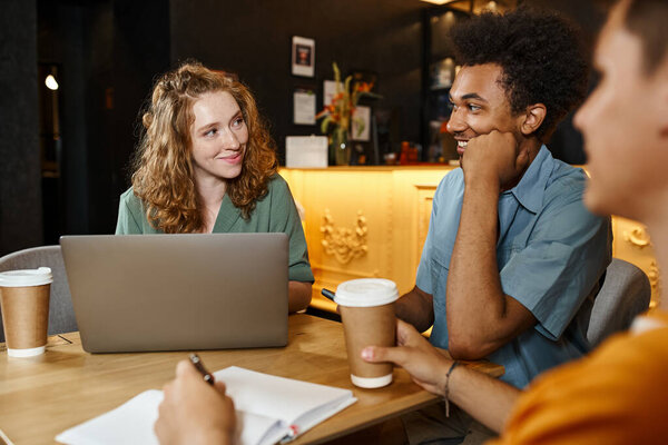 cheerful woman near laptop and multicultural students with paper cup and smartphone in hostel cafe