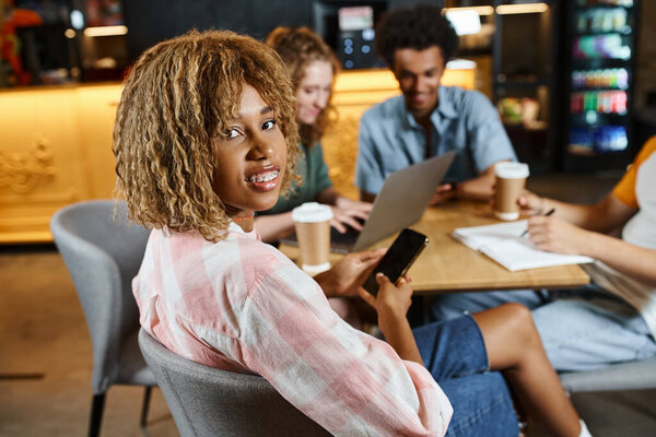 african american woman with smartphone smiling at camera near friends with laptop in hostel cafe