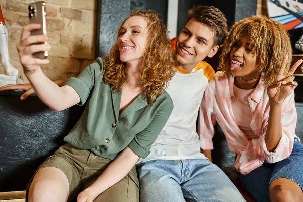 stock image cheerful woman taking selfie with multiethnic friends while having fun in lobby of youth hostel