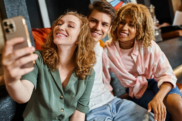 excited woman taking selfie with multiethnic buddies while having fun in lobby of students hostel