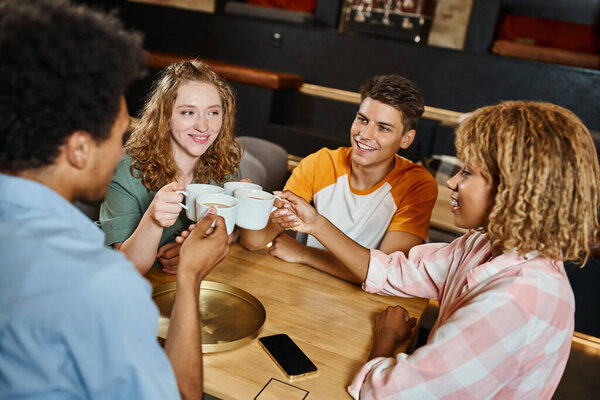 carefree multiethnic students clinking coffee cups in lobby cafe of youth hostel, travelers