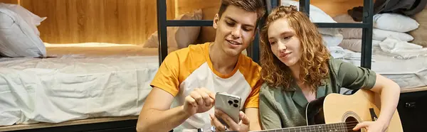 stock image youthful man showing smartphone to girlfriend playing guitar in students hostel, horizontal banner