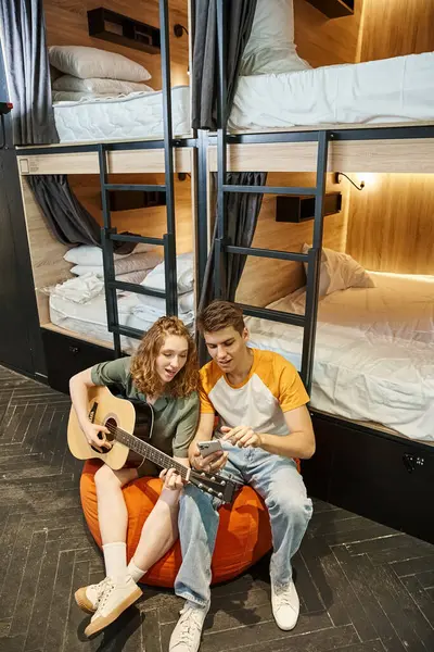 young couple of students with smartphone and guitar sitting on bean bag chair in cozy hostel