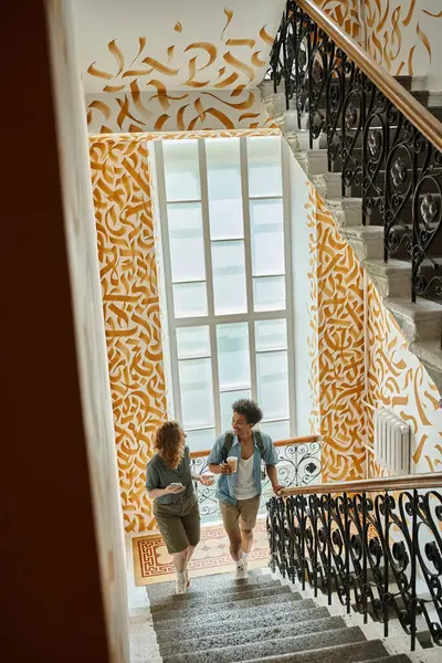 stock image interracial couple with smartphone and paper cup walking on staircase with colorful walls in hostel