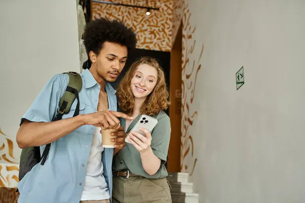 smiling african american man pointing at smartphone near cheerful girlfriend on staircase of hostel