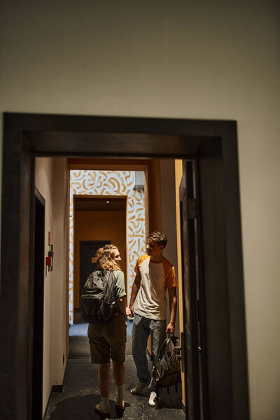 Молодая пара с рюкзаками, держась за руки и разговаривая в коридоре современного студенческого общежития