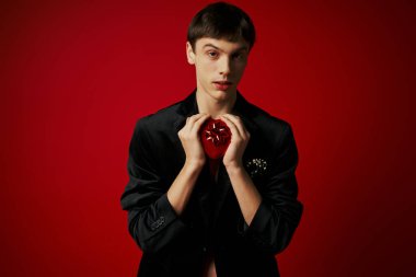 romantic guy holding heart-shaped present and looking at camera on red background, Valentines day clipart