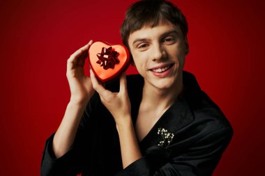 cheerful young man in velvet blazer holding heart-shaped present on red background, Valentines day clipart