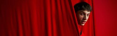 playful young man in vibrant shirt hiding behind red curtain while playing hide and seek, banner clipart