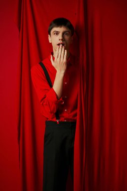 surprised young man in suspenders smiling and covering mouth near red vibrant curtain, merriment clipart